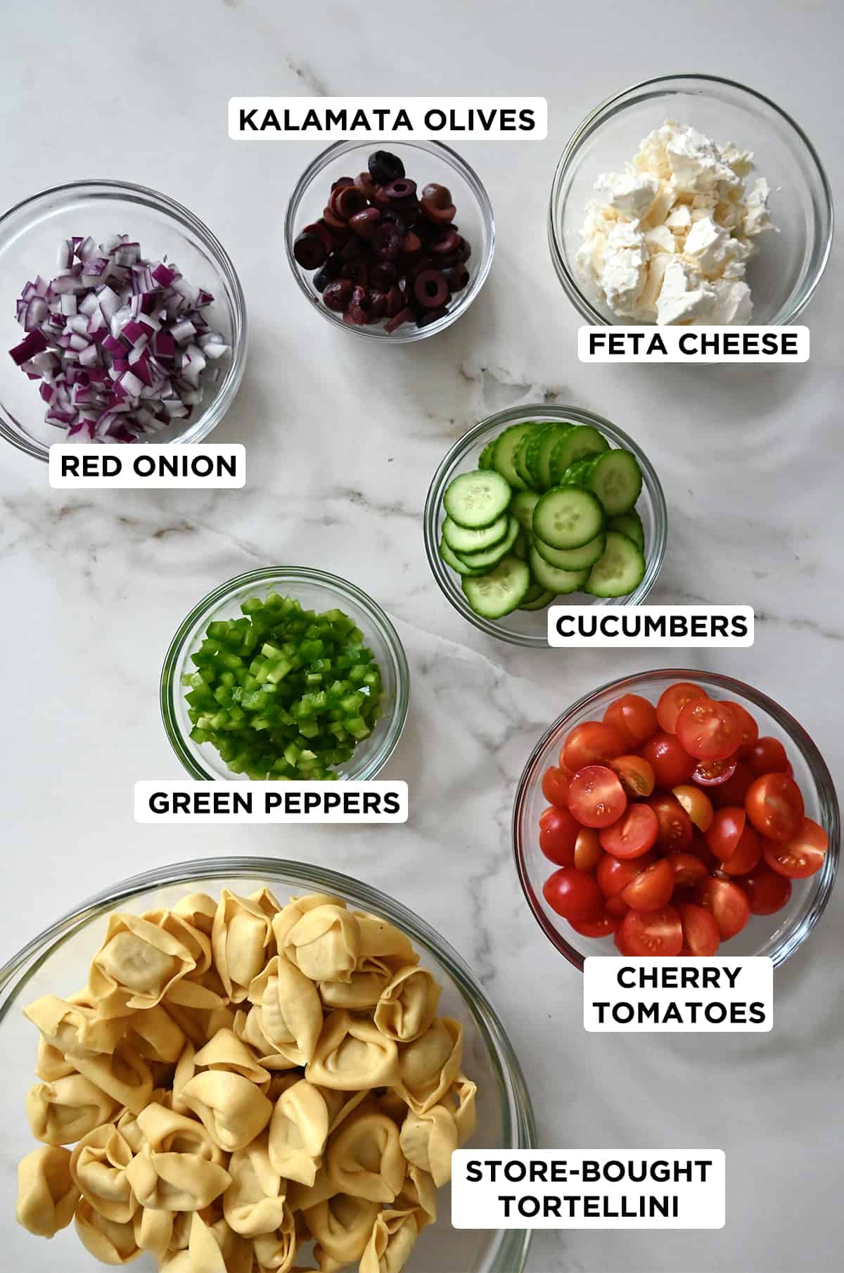 Various sizes of clear bowls containing ingredients for tortellini pasta salad, including feta cheese, cucumber, cherry tomatoes, tortellini, green pepper, red onion and black olives. 