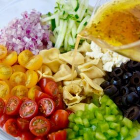 Dressing being poured into bowl containing Greek Tortellini Pasta Salad ingredients