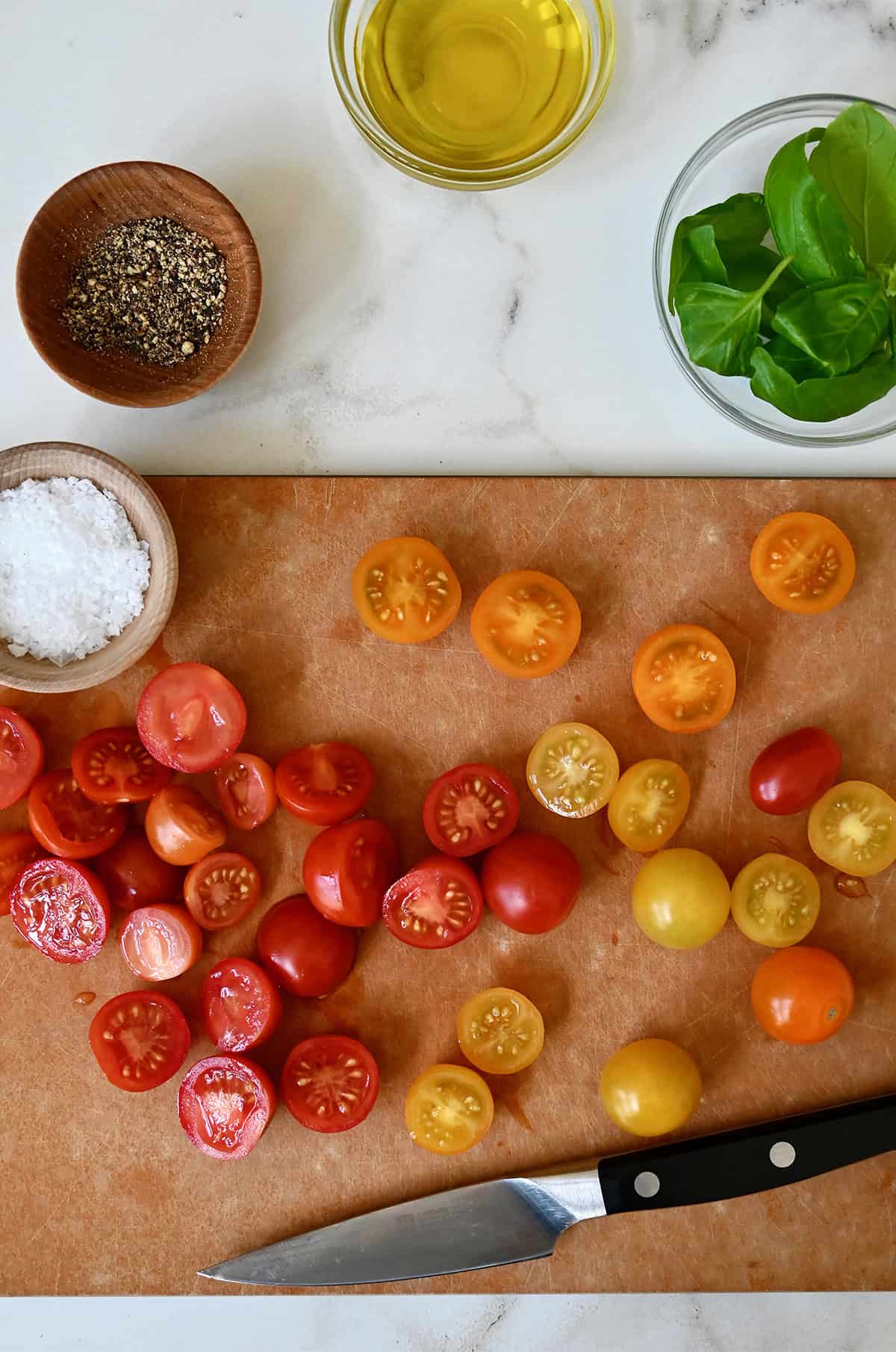 Halved cherry tomatoes on a cutting board with small bowls containing salt, pepper, olive oil and fresh basil leaves nearby.