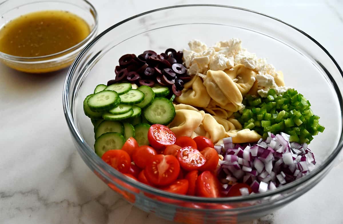 A clear bowl containing tomatoes, cucumbers, olives, feta cheese, cooked pasta, green pepper and red onion next to a small bowl containing red wine vinaigrette.