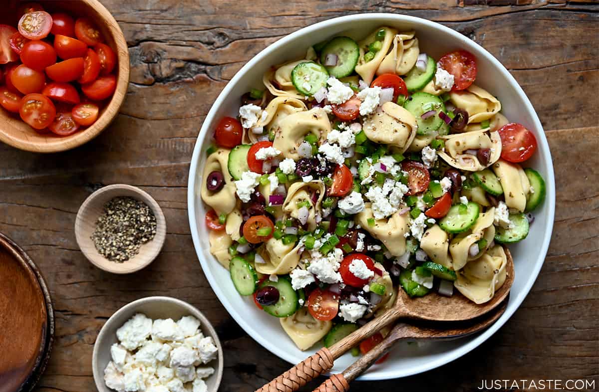 A top-down view of tortellini pasta salad with cherry tomatoes, feta cheese and Kalamata olives in a large white bowl with wooden serving spoons.