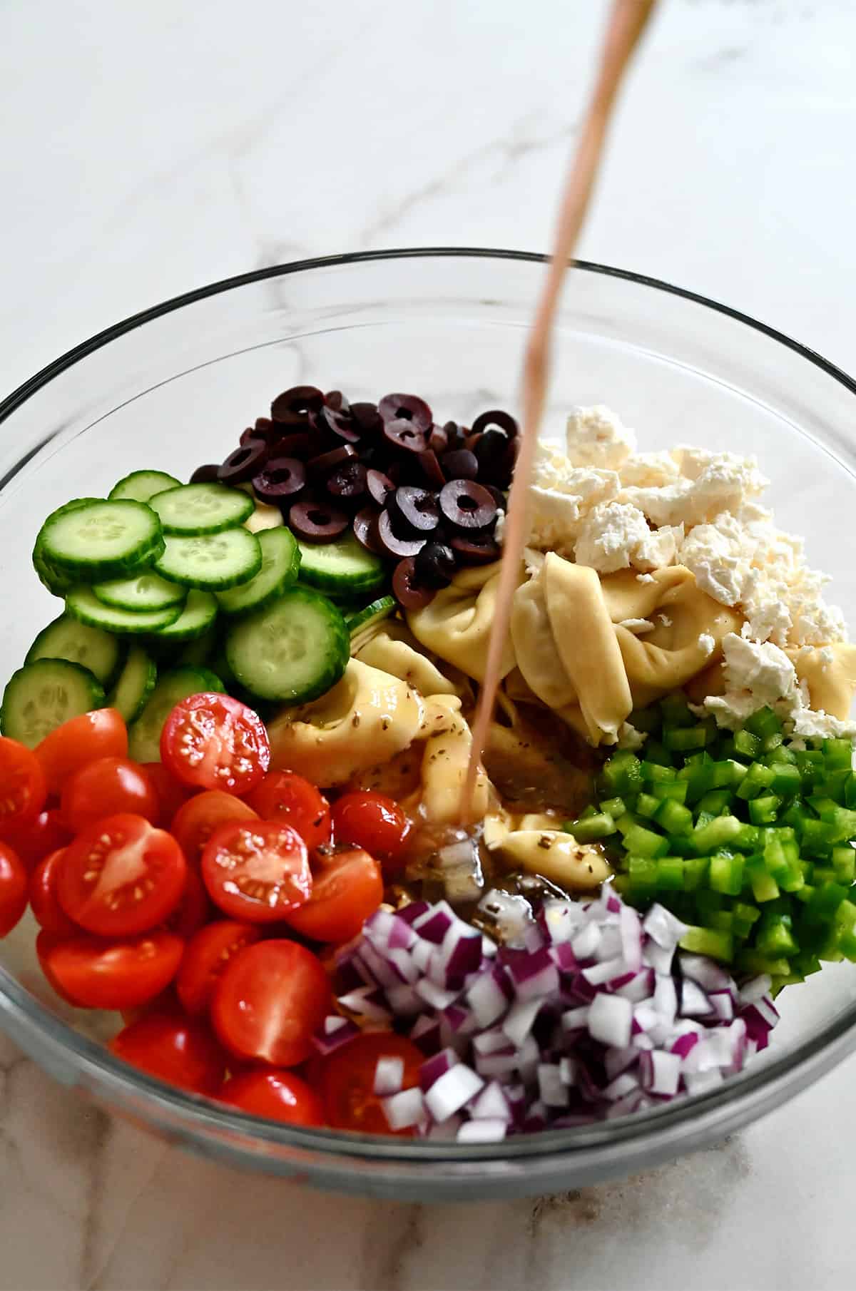 Vinaigrette being poured over halved cherry tomatoes, sliced cucumbers, olives, feta cheese, tortellini, and diced green pepper and red onion in a large clear bowl.