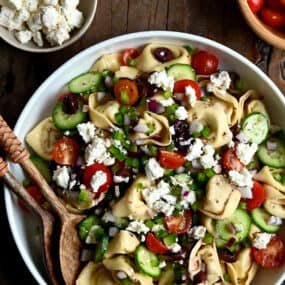 A large serving bowl containing Greek Tortellini Pasta Salad with olives, cherry tomatoes and feta cheese.