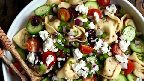 A large serving bowl containing Greek Tortellini Pasta Salad with olives, cherry tomatoes and feta cheese.