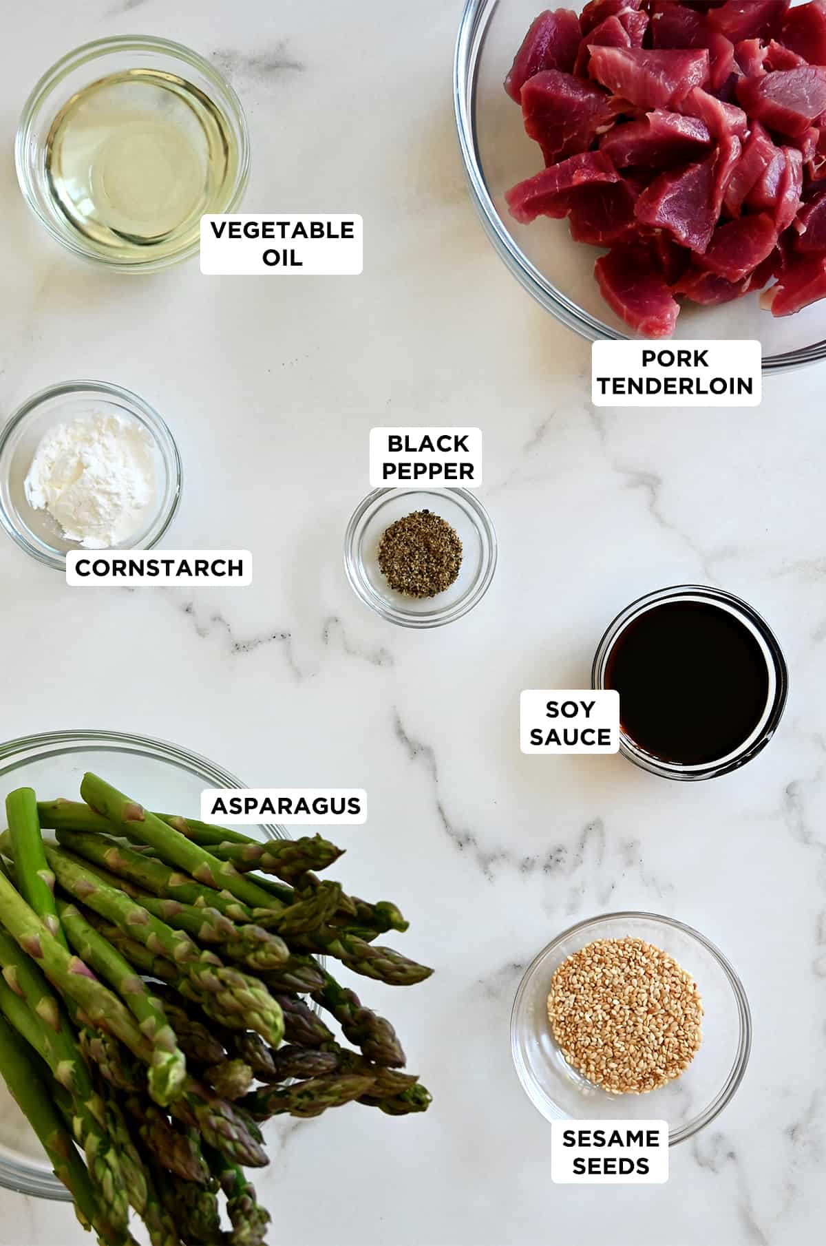 Various sizes of glass bowls containing pork tenderloin, vegetable oil, black pepper, cornstarch, soy sauce, asparagus and sesame seeds.