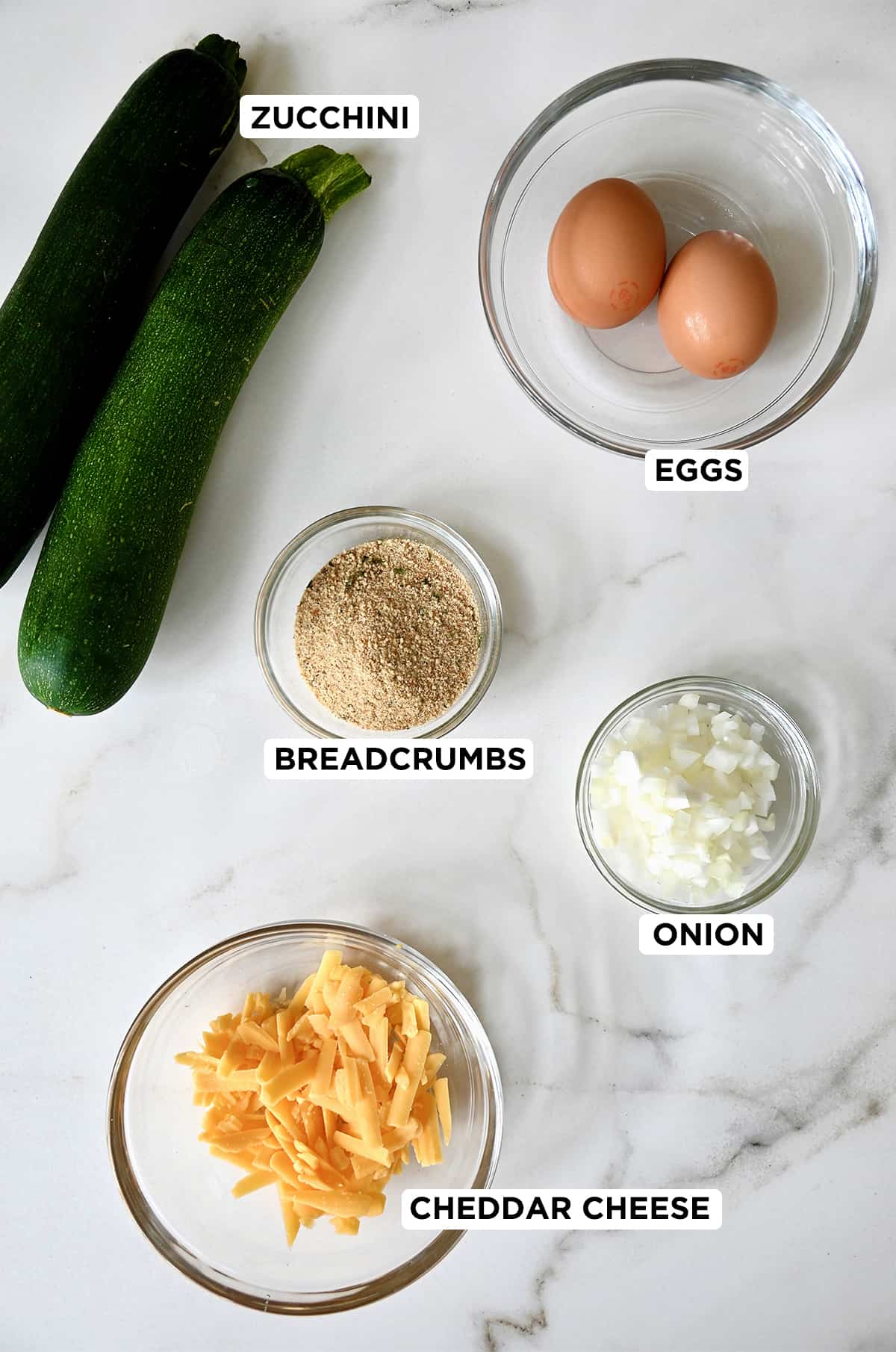 Two zucchinis next to various sized of clear bowls containing eggs, diced onion, breadcrumbs and shredded cheddar cheese.