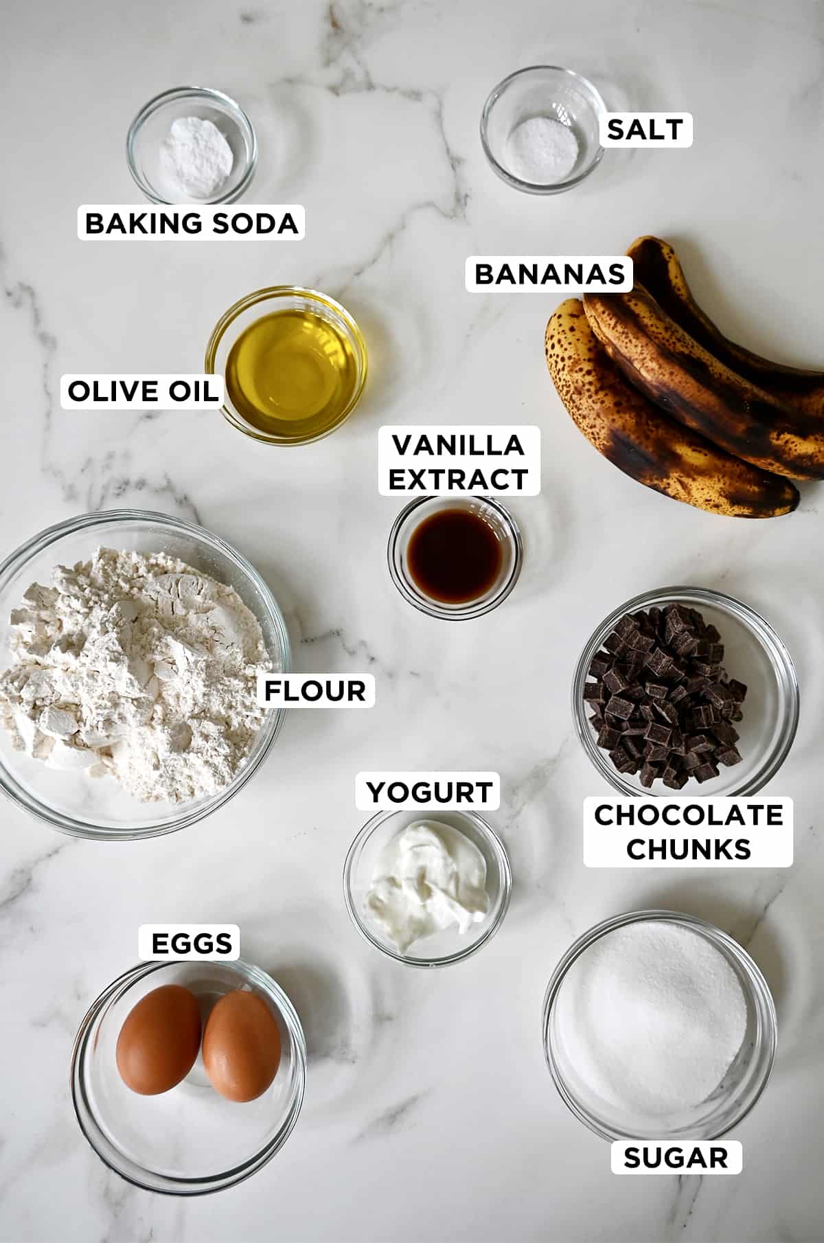 All of the ingredients needed to make one-bowl banana bread, including baking soda, overripe bananas, olive oil, vanilla extract, chocolate chunks, yogurt, flour, eggs, sugar and salt.