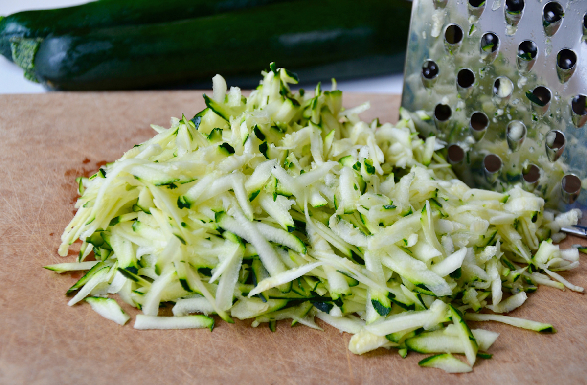 A pile of shredded zucchini on a cutting board next to a box grater.