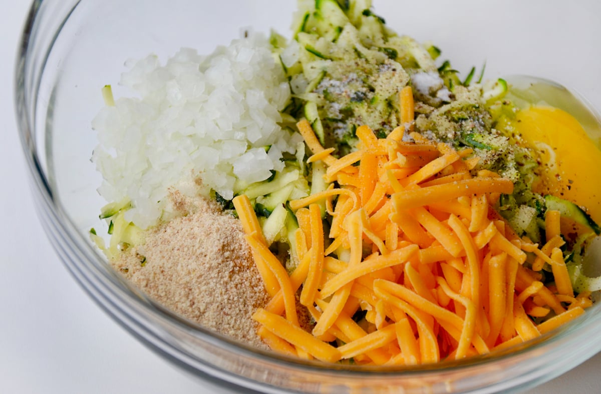 A clear bowl containing diced white onion, shredded zucchini, two freshly cracked eggs, shredded cheddar cheese and breadcrumbs.