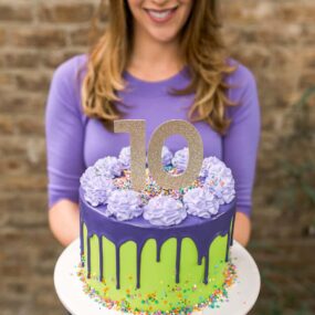 A purple and green drip cake with sprinkles and a girl in the background holding it