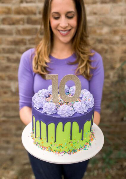 A purple and green drip cake with sprinkles and a girl in the background holding it