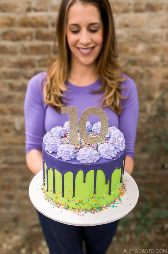 A purple and green drip cake with sprinkles and a girl in the background holding it