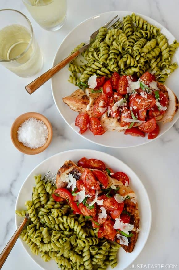 Two plates containing Quick Bruschetta Chicken and pesto pasta next to glasses filled with wine