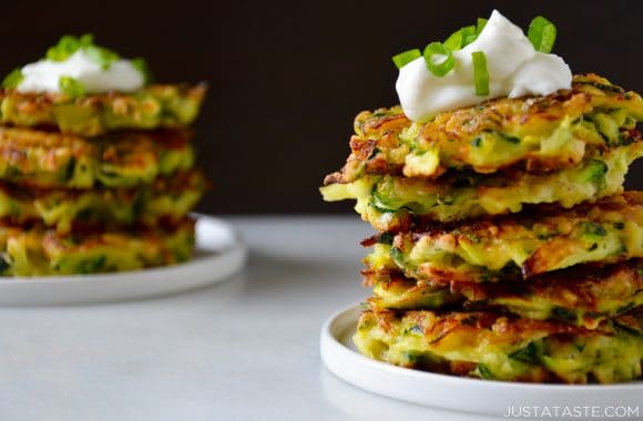 Two white plates containing stacks of zucchini fritters topped with sour cream