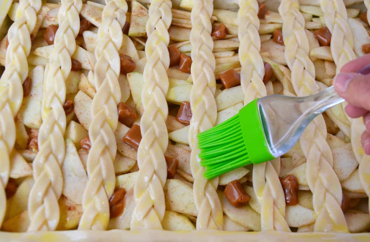 A hand using a pastry brush to brush egg wash onto the braided lattice pie crust before baking.