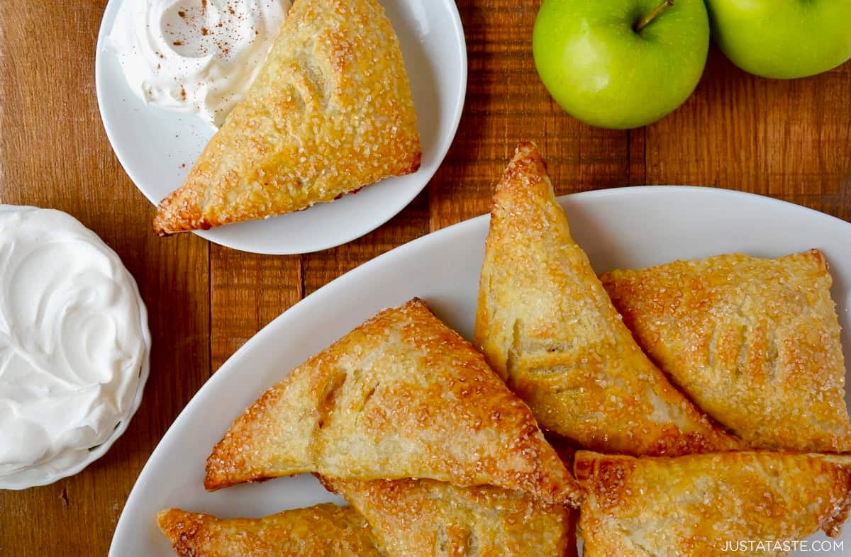 Golden, flaky puff pastry apple turnovers filled with cinnamon apple pie filling on a white serving plate with whipped cream. Two Granny Smith apples are nearby.