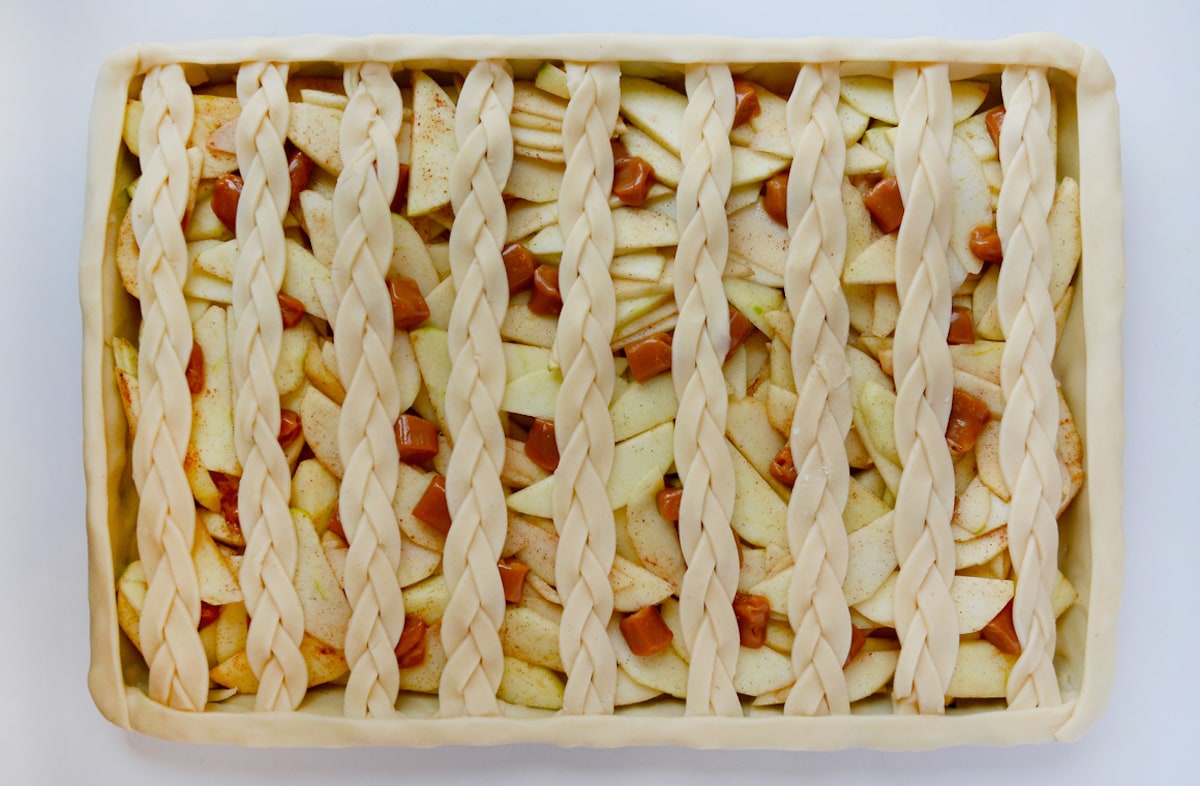 A sheet pan with a fully assembled unbaked pie with a bottom crust, apple filling, soft caramels and braided lattice top crust.