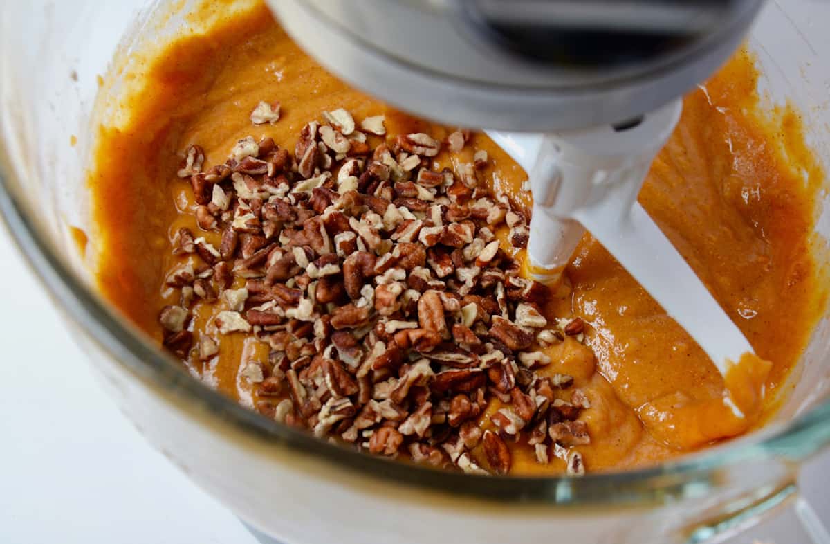 Pumpkin bar batter is in the bowl of a stand mixer, with pecans sprinkled on top, ready to be mixed in.