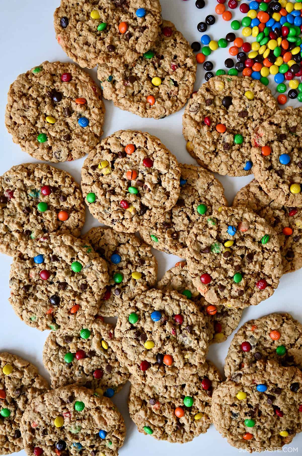 Monster cookies loaded with oats, peanut butter, mini chocolate chips and M&Ms on a white marble background with a handful of M&M candies nearby.