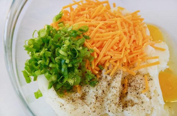 Leftover mashed potatoes, chopped scallions, shredded cheddar cheese and eggs in clear bowl