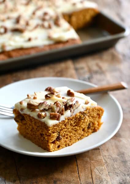 A slice of pumpkin bar topped with cream cheese frosting and toasted pecans on a plate with a fork.