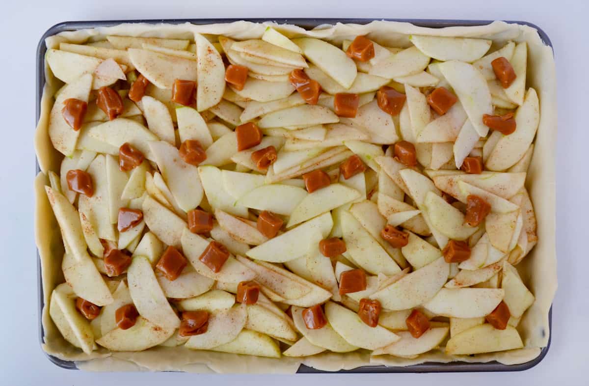 A sheet pan containing pie dough topped with apple slices, soft caramels and ground cinnamon.