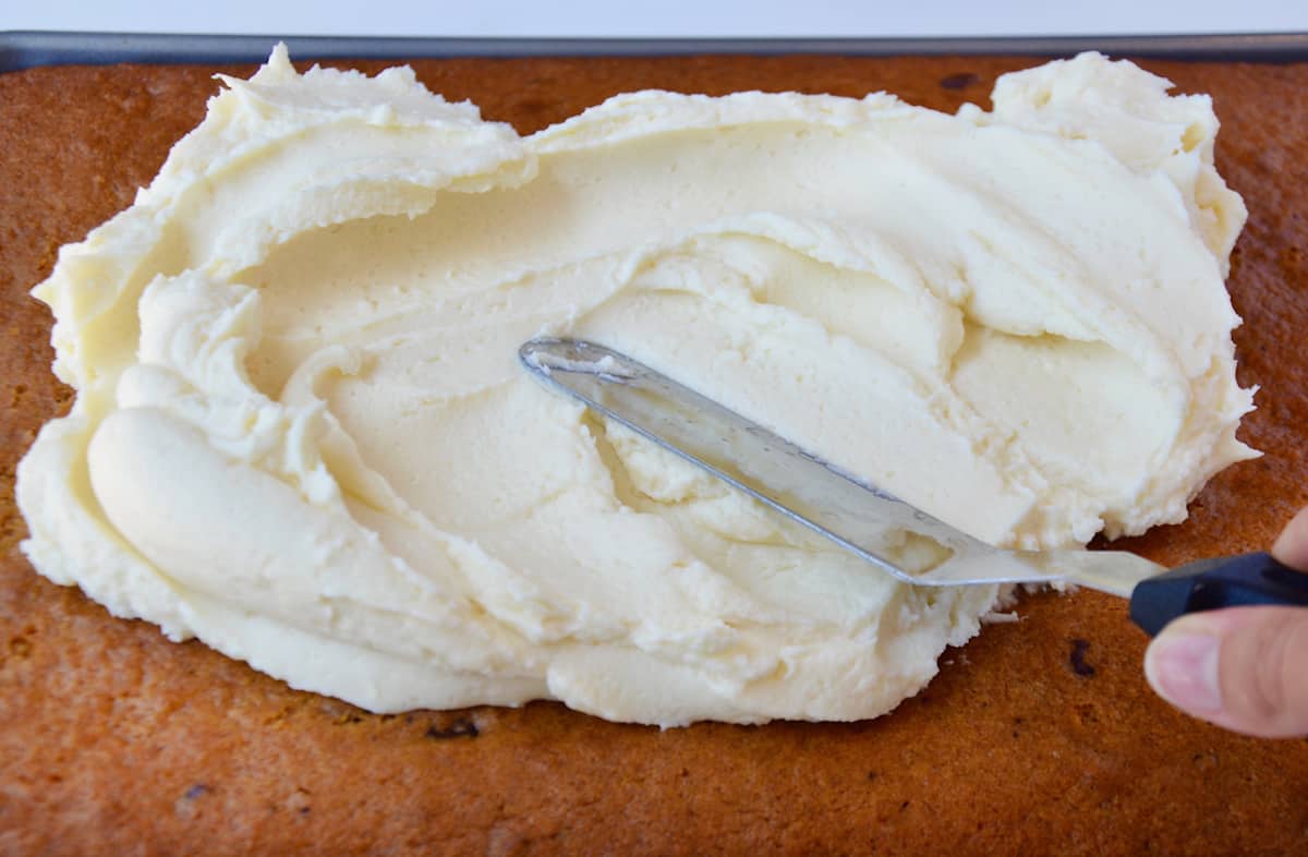 An offset spatula is being used to spread cream cheese frosting on a pan of pumpkin bars.