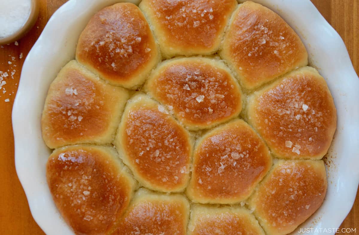 Golden brown dinner rolls topped with large-flake sea salt in a white baking dish.