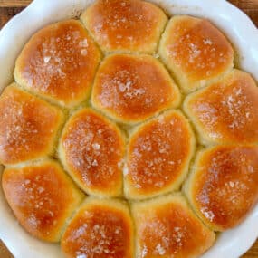 Freshly baked soft dinner rolls garnished with large-flake sea salt in a white round baking dish.