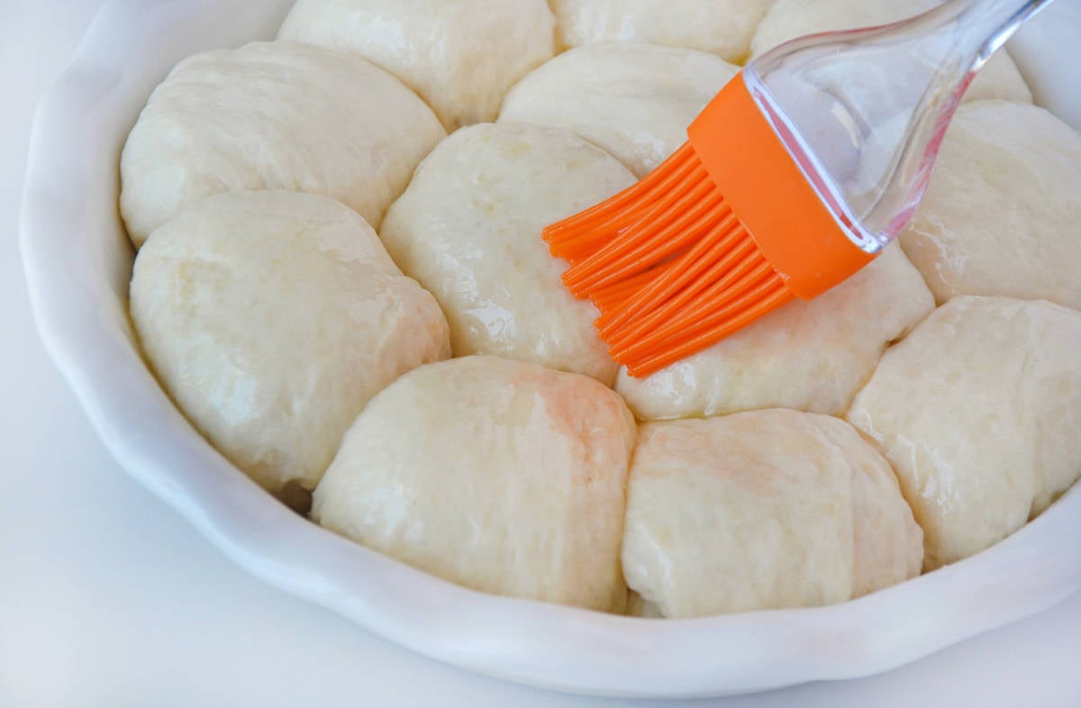 A pastry brush brushes butter atop unbaked dinner rolls in a white baking dish.