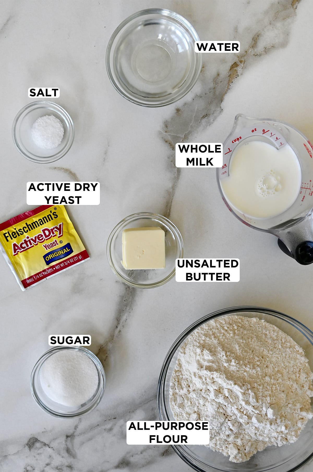 A packet of active dry yeast and a liquid measuring cup filled with whole milk are next to various sizes of clear bowls containing ingredients for dinner rolls, including water, butter, flour, sugar and salt.