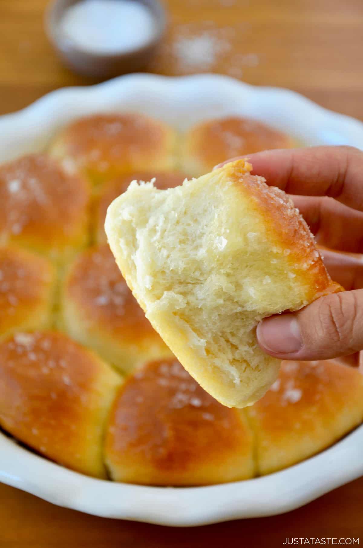 A hand holds a soft dinner roll above a baking dish containing homemade rolls.