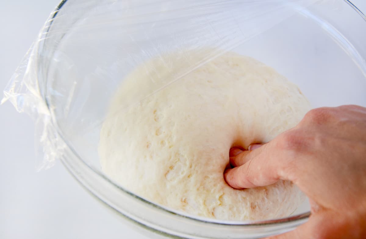 A hand presses into bread dough in a clear bowl that's half covered with plastic wrap.