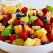 Orange poppy seed dressing being poured over fresh fruit in a large white serving bowl.