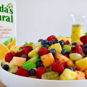 A white bowl with fruit salad and a carton of orange juice in the back