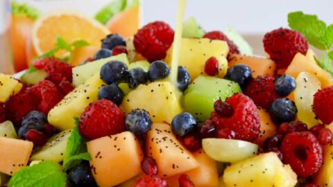 A white bowl with fruit salad with orange poppy seed dressing being poured on top