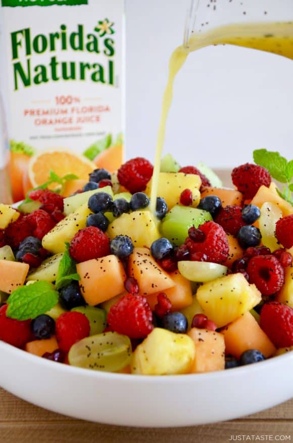 A white bowl with fruit salad with orange poppy seed dressing being poured on top