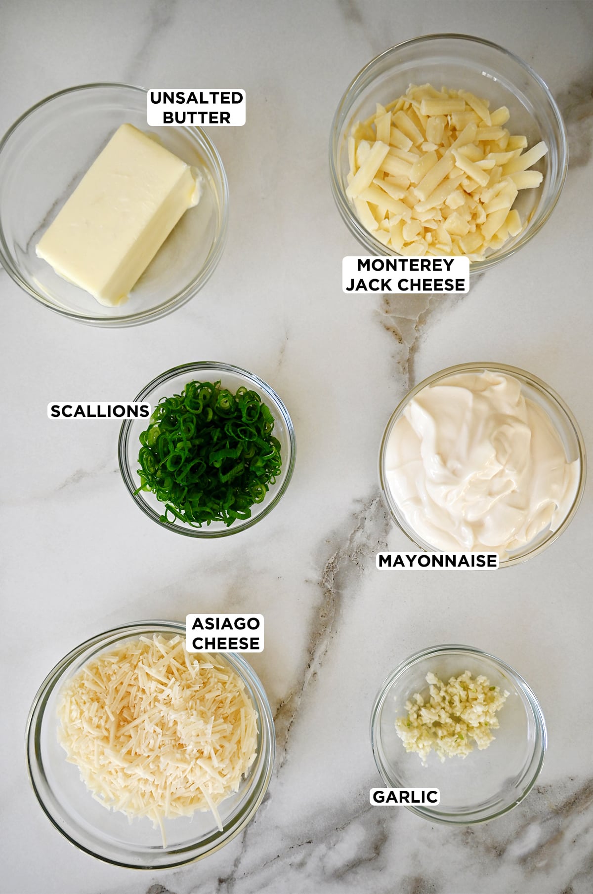 Glass bowls containing cheesy garlic bread ingredients, including butter, Monterey Jack cheese, mayonnaise, scallions, asiago cheese and minced garlic.