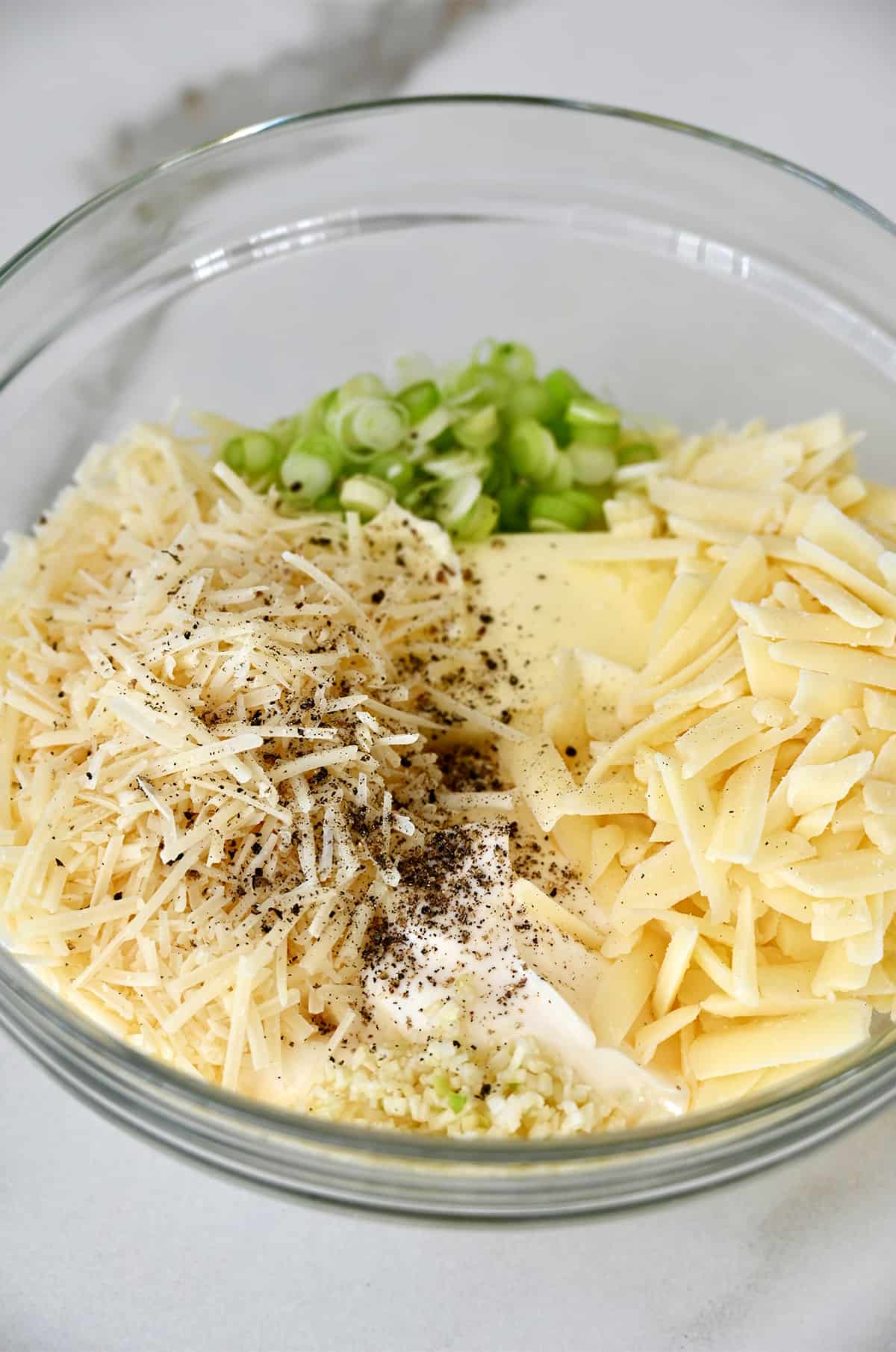 A glass bowl containing butter, shredded asiago and Monterey Jack cheese, sliced scallions, mayonnaise, salt and pepper.