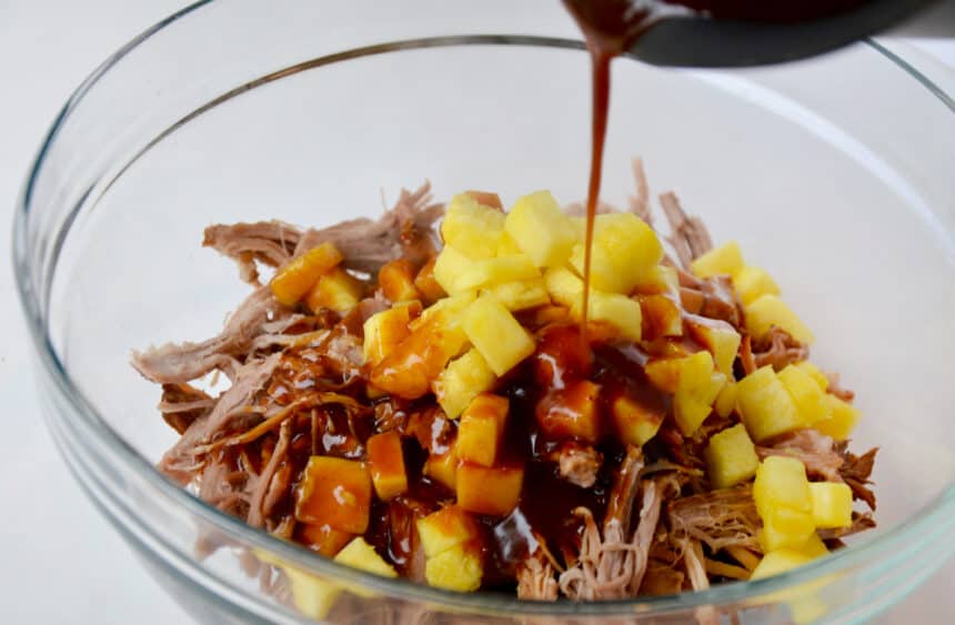 Barbecue sauce being poured over pineapple and shredded pork in a clear bowl