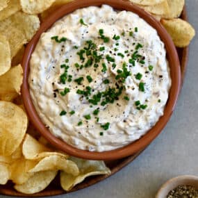 Sour cream and onion dip topped with fresh chives and black pepper in a small bowl on a plate with classic potato chips.
