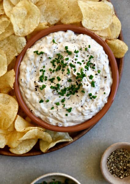 Sour cream and onion dip topped with fresh chives and black pepper in a small bowl on a plate with classic potato chips.