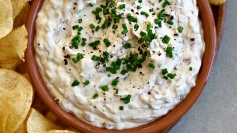 Sour cream and onion dip topped with fresh chives and black pepper in a small bowl on a plate with classic potato chips.