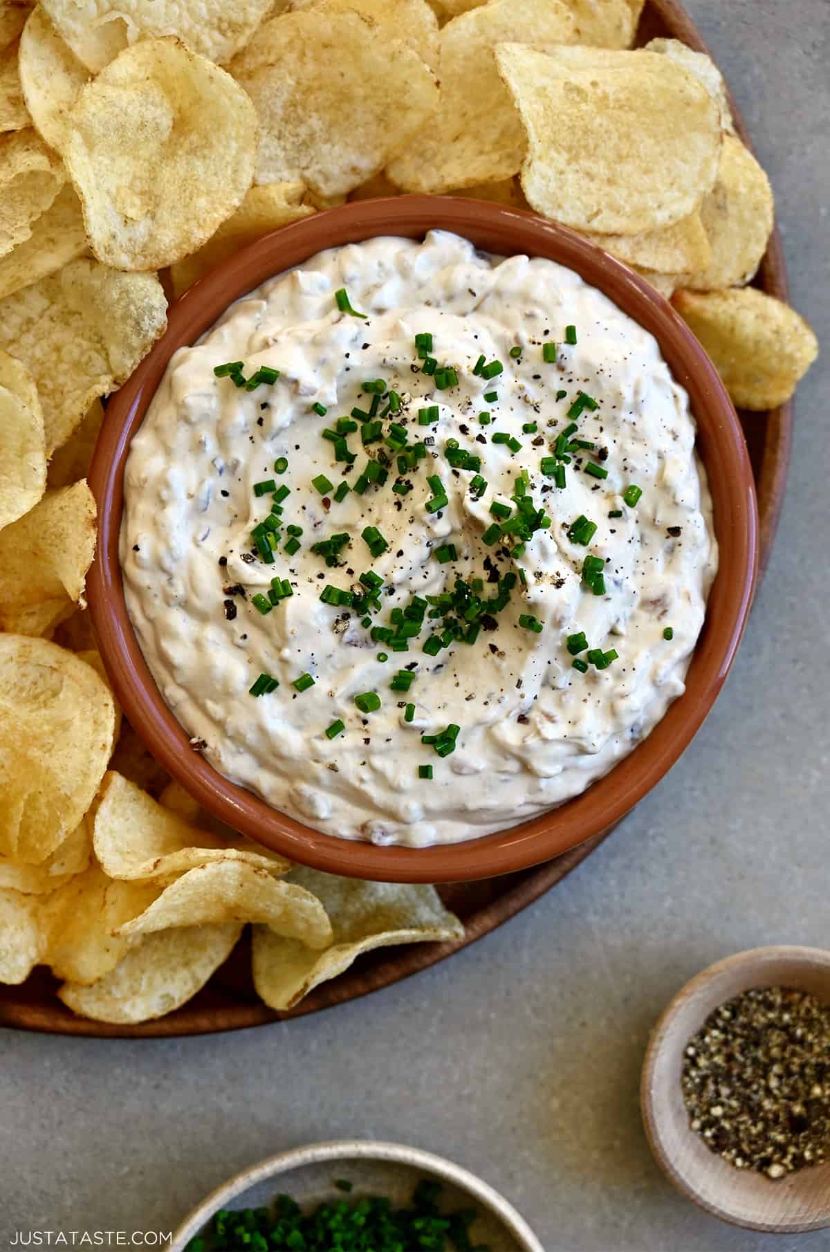 Sour cream and onion dip topped with fresh chives and black pepper in a small bowl on a plate with classic potato chips.