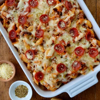 Top-down view of Pepperoni Pizza Pasta in white baking dish on wood board next to slices of Italian bread and small ramekins with parmesan cheese and black pepper