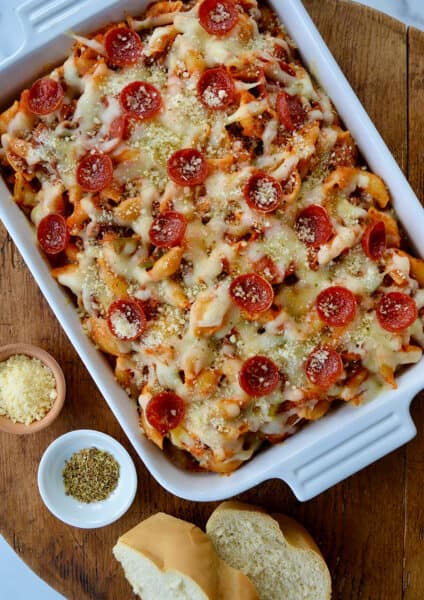 Top-down view of Pepperoni Pizza Pasta in white baking dish on wood board next to slices of Italian bread and small ramekins with parmesan cheese and black pepper