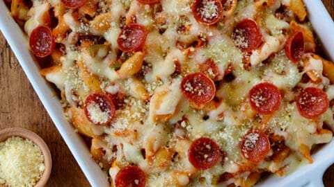 Top-down view of Pepperoni Pizza Pasta in white baking dish on wood board next to slices of Italian bread and small ramekins with parmesan cheese and black pepper