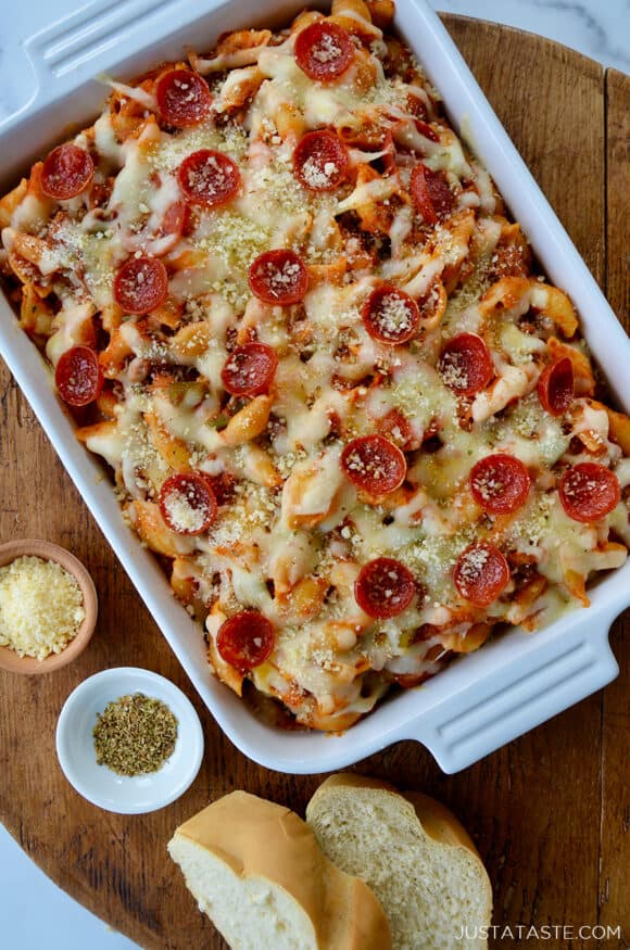 Top-down view of Pepperoni Pizza Pasta in white baking dish on wood board next to slices of Italian bread and small ramekins with parmesan cheese and black pepper