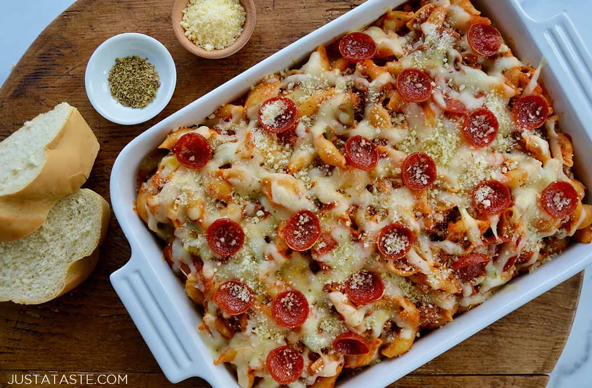 Top-down view of homemade Pepperoni Pizza Pasta in white baking dish next to two slices of Italian bread and small bowls containing Parmesan cheese and black pepper