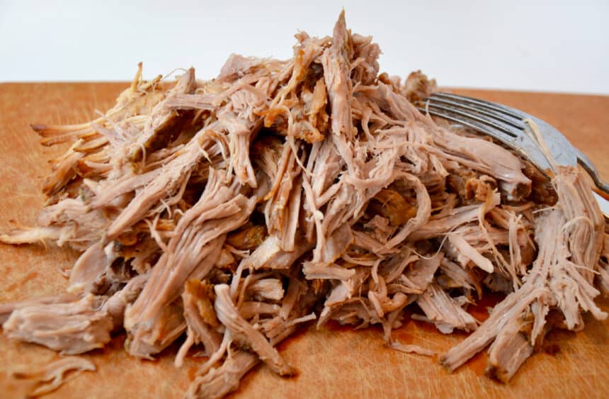 A pile of pulled pork on a cutting board next to a fork